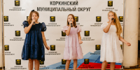 На фото три девочки в нарядных платьях выступают с микрофонами на фоне пресс‑волла с символикой Коркинского муниципального округа. - Коркинский муниципальный округ