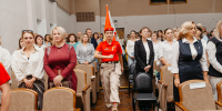 В зале с рядами кресел молодой человек в красной футболке, бежевых брюках и оранжевом головном уборе с флагом проходит между рядами, по обе стороны от него стоят и сидят участники мероприятия — преимущественно женщины в деловой одежде. - Коркинский муниципальный округ
