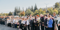 На фото — группа школьников и взрослых, выстроившихся в линию на улице: дети в парадной форме с цветами и медалями, рядом — учителя и другие взрослые, на фоне — городские здания и деревья. - Коркинский муниципальный округ