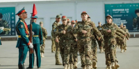 На фото — строевой смотр: колонна военнослужащих в камуфляже марширует по плацу, слева стоит офицер в парадной форме с винтовкой и красным флажком, на фоне — здание с информационными баннерами. - Коркинский муниципальный округ