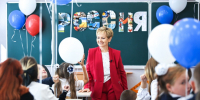 Улыбающаяся учительница в ярко‑красном костюме в классе среди детей в школьной форме с воздушными шарами; на доске видна надпись «РОССИЯ», составленная из фотоколлажей. - Министерство образования и науки
