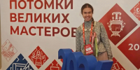 Улыбающаяся девушка в народном стиле на фоне баннера Всероссийского фестиваля «Потомки великих мастеров»: перед ней объёмные синие цифры «2026» на красной подставке, а на фоне — орнаменты и символ самовара. - Министерство образования и науки