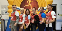 Шестеро взрослых в красных жилетах ГТО с номерами на груди стоят в ряд на фоне большой эмблемы/баннера фестиваля, показывают «палец вверх». - 168.Ru