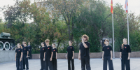 На фото — группа молодых людей в чёрных костюмах с красными звёздами на груди стоит на открытой площадке у мемориала; один из них держит микрофон, на фоне — танк, деревья и флаги. - Коркинский муниципальный округ