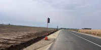 На фото уходящая вдаль дорога. Навстречу наблюдателю едет белый автомобиль. Справа высохшее кукурузное поле. Слева равнина и песчаная обочина со следом протектора - Министерство транспорта