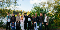 Группа молодых людей и взрослых на берегу реки: некоторые держат распечатки/дипломы, у части — сумки/пакеты; на заднем плане видна вода и прибрежная растительность. - 168.Ru