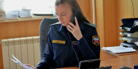 Сотрудница в форме ФССП России за рабочим столом держит телефон и просматривает бумаги. - 168.Ru