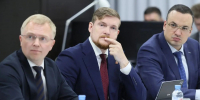 На изображении трое мужчин в деловых костюмах сидят за столом. На столе ноутбук, бутылки с водой и таблички. Мужчины выглядят сосредоточенными., Изображение. - Минэкономразвития