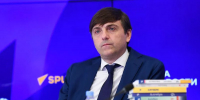 мужчина в тёмном костюме сидит за столом на фоне баннера «РИА Новости», перед ним несколько стопок учебников и микрофон - 168.Ru