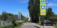 Узкая улица с дорожными знаками перехода, неровности и ограничения скорости; справа припаркован автомобиль, вдали церковь - 168.Ru