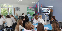В учебном помещении проходит занятие: группа подростков обсуждает материал, а преподаватель демонстрирует на экране видеоконференцию, в помещении также видны флаги и информационный стенд. - Министерство образования и науки