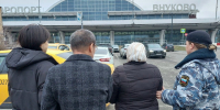Сотрудница ФССП ведёт задержанную женщину и ещё несколько человек на парковке на фоне здания аэропорта с надписью «ВНУКОВО» - 168.Ru