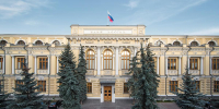 Фасад здания Банка России с российским флагом на крыше и хвойными деревьями на переднем плане - 168.Ru