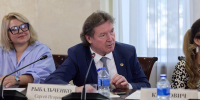 Мужчина в костюме за столом на совещании; перед ним микрофон, бутылки с водой и настольная табличка с фамилией. - 168.Ru
