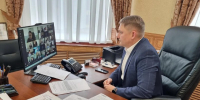мужчина в офисе за столом смотрит на экран с сеткой участников видеоконференции; на столе документы, телефон и оргтехника - 168.Ru