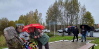 Люди под зонтами возлагают венки к памятному камню на мощёной площадке; на заднем плане дорога, автомобили и осенние деревья. - 168.Ru