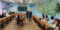 Учебный класс: школьники сидят за партами, два пожилых мужчины (ветераны) стоят у переднего стола с предметами из экспозиции (фуражка, каска, атрибуты), на столах — листовки о полиции. - 168.Ru