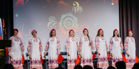На сцене — женский ансамбль в традиционных русских костюмах с красно‑белой вышивкой, в руках участниц — красные платки и колокольчики, на фоне — декоративный орнамент; перед сценой — силуэты зрителей. - Коркинский муниципальный округ