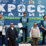 Призёры забега на пьедестале перед большим баннером «Кросс нации»: спортсмены с медалями и дипломами на открытой сцене в лесопарковой зоне. - 168.Ru
