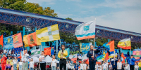 На спортивном стадионе проходит массовое мероприятие: группа людей с флагами муниципальных образований стоит на поле, на фоне — трибуны со зрителями и металлическая конструкция крыши. - Министерство образования и науки
