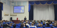 В актовом зале проходит лекция «Финансовое мошенничество: стратегии защиты» — спикер выступает перед аудиторией студентов на фоне презентационного слайда и сценического занавеса. - Министерство образования и науки