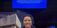 На фото Наталья Михайловна Милова — призёр всероссийских профессиональных конкурсов Минпросвещения РФ 2026 года: она держит диплом и букет цветов на фоне экрана с названием церемонии и стилизованного изображения здания. - Министерство образования и науки