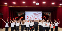 На сцене группа девушек в белых футболках и тёмных брюках участвует в праздничном номере на мероприятии «Посвящение в студенты», размахивая красными флажками на фоне тематического баннера и декораций. - Министерство образования и науки