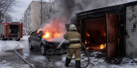 пожарный в спецформе поливает из рукава горящий легковой автомобиль рядом с горящим гаражом на заснечной улице; пожарная машина на заднем плане - 168.Ru