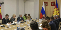 На фото — официальное совещание в конференц‑зале: участники в деловой одежде сидят за длинным столом с табличками, микрофонами и бутылками воды, на фоне видны флаги РФ и субъекта РФ, а также гербы на стене. - Министерство образования и науки