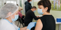 медицинская сестра в маске и шапочке делает укол в плечо взрослой женщины в прививочном кабинете, на заднем плане сидящие в масках люди - 168.Ru