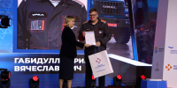 На сцене женщина вручает Ринату Габидуллину сертификат и подарочный пакет на церемонии награждения в рамках конкурса «Лучший по профессии» — на фоне экрана с изображением спецодежды и грузовика «Урал» и табличкой с именем награждаемого. - Коркинский муниципальный округ