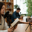 В читальном зале библиотеки девушка читает вслух в микрофон; на столе смартфон на штативе записывает, вокруг книжные полки и другие читатели. - 168.Ru