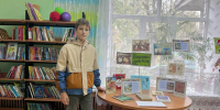 Мальчик стоит в детском отделе библиотеки рядом со столом-выставкой книг и табличкой «Малочисленные народы России», на полках видны детские книги. - 168.Ru