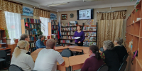 Группа людей в читальном зале библиотеки слушает женщину-ведущую на фоне книжных полок и экрана с биографическими слайдами. - 168.Ru