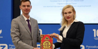 На фото мужчина в сером костюме и женщина в чёрном пиджаке держат книгу «Тайны русских народных сказок» на фоне баннера Российского общества «Знание» и слайда о популяризации духовно‑нравственной культуры в рамках серии «Удивительная Русь». - Министерство образования и науки