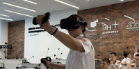 Юноша в VR‑шлеме с контроллерами демонстрирует работу с технологией виртуальной реальности в учебном классе, на фоне — другие учащиеся в форменной одежде и информационные стенды. - Министерство образования и науки