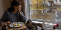 Женщина завтракает у окна после дождя; на столе яичница, а рядом с архивной фотографией Кавказских гор лежат две красные гвоздики, за окном видны отражения деревьев в лужах. - 168.Ru