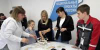 На фото группа взрослых и ребёнок совместно собирают каркасную модель в рамках образовательного мероприятия, на фоне — стенд с надписью «Образование. Национальные проекты России». - Министерство образования и науки
