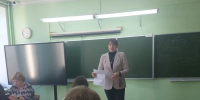 Взрослая женщина стоит у школьной доски и держит раздаточные листы; перед ней – несколько школьников за партами; в классе видны интерактивный экран и мелованная доска. - 168.Ru