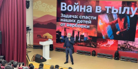 На фото мужчина в деловом костюме выступает на сцене перед аудиторией; на большом экране — слайд с заголовком «Война в тылу. Задача: спасти наших детей от вербовки» и тематическими изображениями. - Министерство образования и науки
