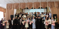 На фото — группа педагогов и священнослужителей на торжественной церемонии награждения: участники держат в руках дипломы и букеты цветов, на фоне — презентационный экран и оформленная сцена. - Министерство образования и науки