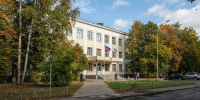 Бело-жёлтое здание поликлиники с флагами у входа среди зелени солнечным днём - Newsroom24.Ru
