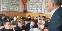 Взрослый инструктор проводит урок по технике безопасности для школьников в классе; на столах — спасательное снаряжение (аптечка, изолирующий коврик/покрытие, дыхательные средства), на стенах — плакаты по защите и спасению. - 168.Ru