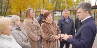 Мужчина беседует с группой жителей на вымощенной плиткой аллее сквера; на заднем плане лавочки, навесы и деревья с осенней листвой. - 168.Ru