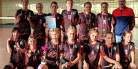 Девочки‑волейболистки в игровой форме с медалями, кубком и грамотой на фоне волейбольной сетки в спортивном зале, рядом взрослый тренер - 168.Ru