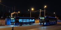Два синих электробуса с подсветкой стоят на конечной остановке вечером под фонарями - Newsroom24.Ru