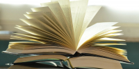 Раскрытая книга с веером страниц поверх стопки других книг на столе - Newsroom24.Ru