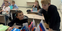 Младшие школьники в классе за партами во время занятия; на переднем плане учебники и распечатки, одна из которых — иллюстрация с ребёнком и мобильным телефоном/монстром. - 168.Ru