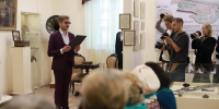 мужчина в костюме читает с папкой в экспозиционном зале музея; перед ним сидящая аудитория, справа — фотографы и музейная карта на стене - 168.Ru