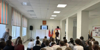 Школьное собрание в актовом/малом зале: ряд учащихся, сидящих лицом к ведущей у экрана; на стене герб, рядом флаги. - 168.Ru